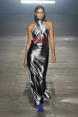 Dark-haired model wearing a metallic dress with red writing on a runway